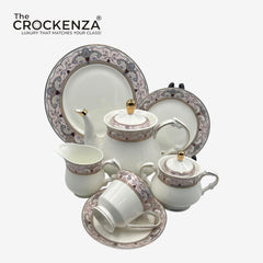 A crockeware tea and coffee set from The Crockenza, featuring a white porcelain teapot, sugar bowl, creamer, cups, plates, and lids decorated with a purple and gold floral border pattern, set against a light gray background.