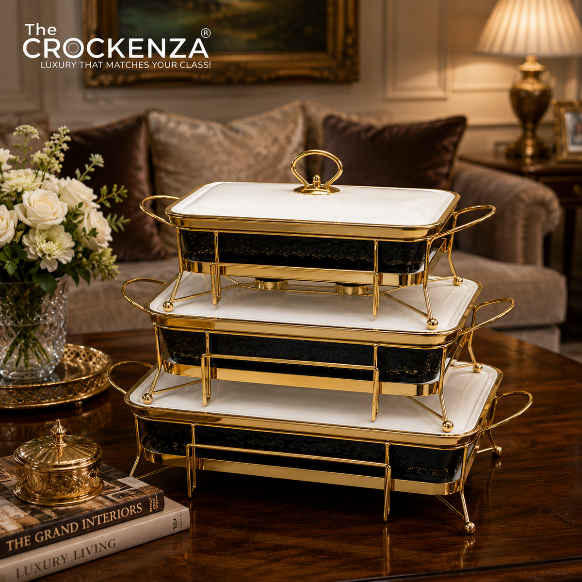 Three-tiered rectangular chafing dishes with white marble-look tops, black marble-look sides, and gold metal frames, displayed on a wooden table in a luxurious living room setting. The brand logo 'The CROCKENDA' is visible in the top left corner.