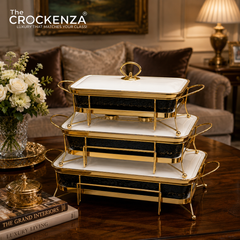 Three-tiered rectangular chafing dishes with white marble-look tops, black marble-look sides, and gold metal frames, displayed on a wooden table in a luxurious living room setting. The brand logo 'The CROCKENDA' is visible in the top left corner.