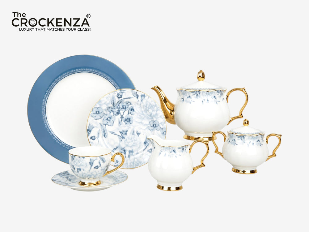 The Crockenza luxury tea and coffee set in white porcelain with blue floral patterns and gold accents, featuring a teapot, sugar bowl, creamer, teacup, and saucer, alongside a blue-rimmed plate, on a light gray background with the Crockenza logo in the top left corner.