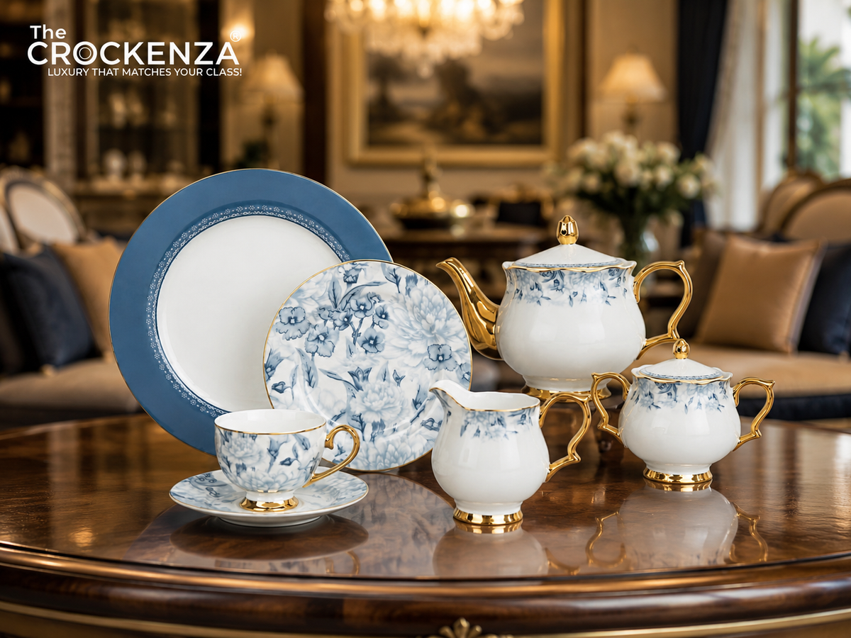 A Crockenza porcelain tea and coffee set with blue floral patterns and gold trim, displayed on a polished wooden table in a luxurious living room setting. The set includes a teapot, creamer, sugar bowl, teacup and saucer, a large plate, and a smaller plate, with 'The CROCKENZA' logo in the top left corner.