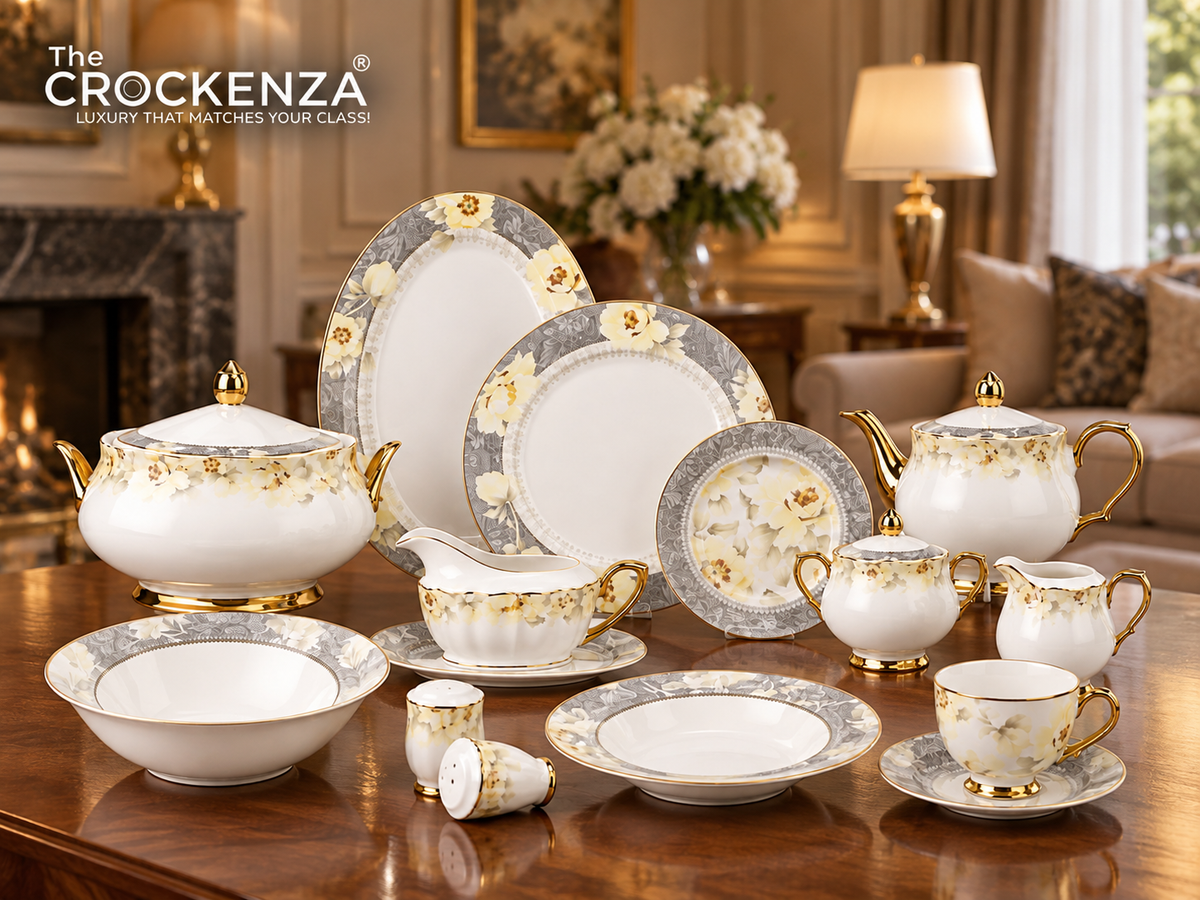 A Crockenza porcelain tea and coffee set arranged on a polished wooden table in a luxurious living room setting. The set includes a teapot, teacups with saucers, a sugar bowl and holder, a creamer, a bowl, plates, and small dishes, all decorated with elegant gold floral patterns and gold trim on a white base.